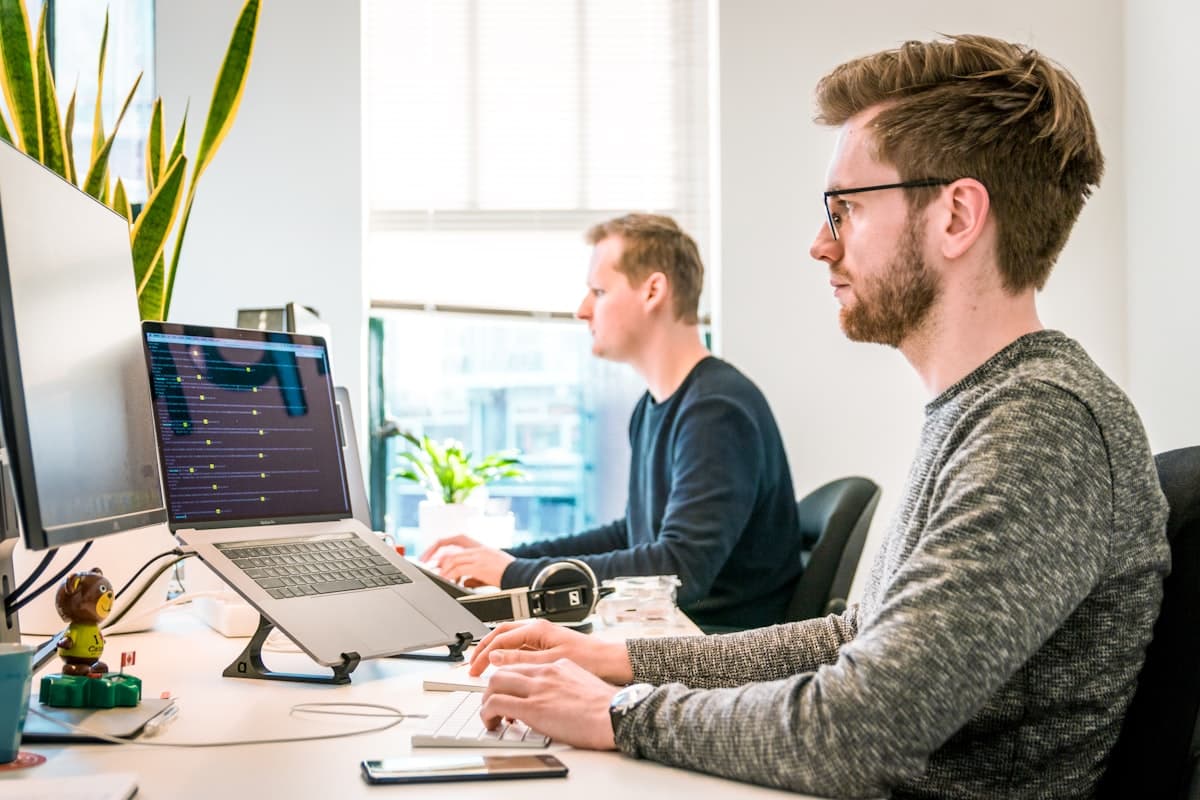Two colleagues at desks with a monitor and a laptop in a bright office